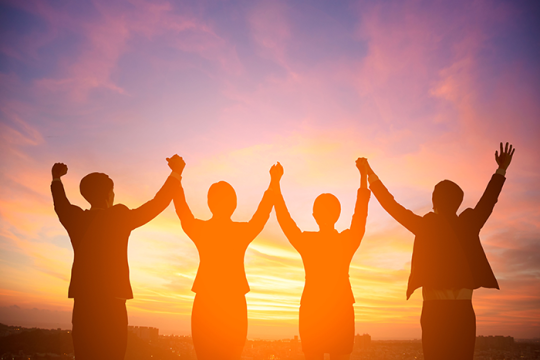 Photo of four people's silhouettes holding hands up in the air; with the sun setting in front of them