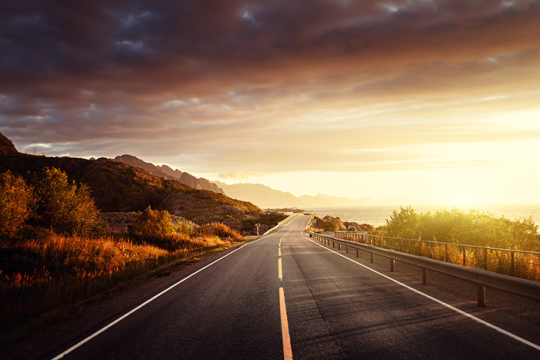 Photo of a road with hills on one side, the ocean on the other and the sut setting in the background