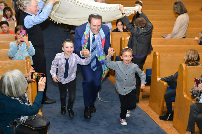 Rabbi Michael Dolgin holds hands with two young children beneath a prayer shawl in the middle of synagogue pews