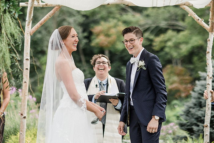 an image of two people of the same sex, one wearing a dress and one wearing a suit, under the chuppah getting married