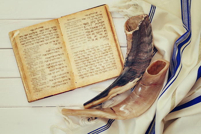 an image of a prayer book opened, two shofars and a tallit on a white wooden surface