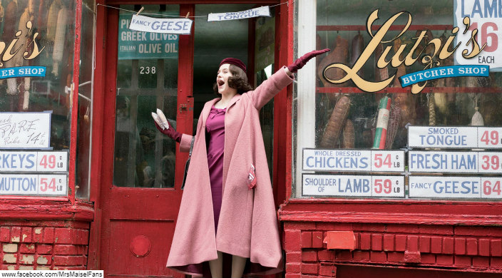Young woman in a red velvet hat and a pink overcoat flings her arm above her head as though singing an expressive song in front of an old fashioned butcher shop