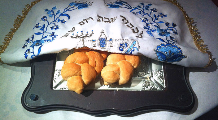 Two braided Shabbat challot on a tray beneath a white and gold embroidered challah cover