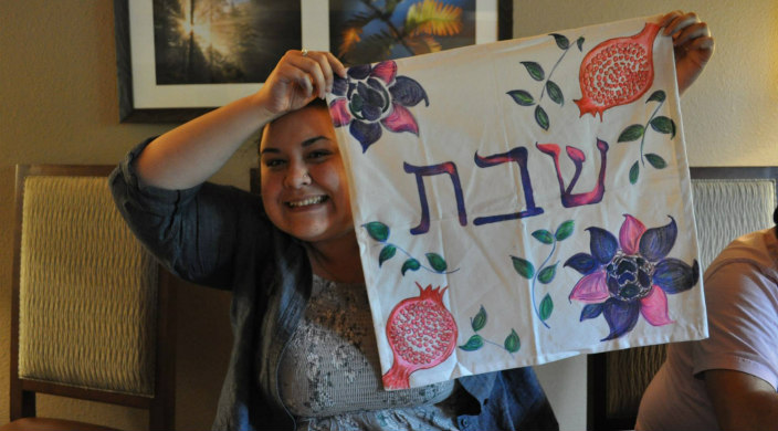 Amanda Ryan smiles while holding up a challah cover with Hebrew words and a hand drawn pomegranate