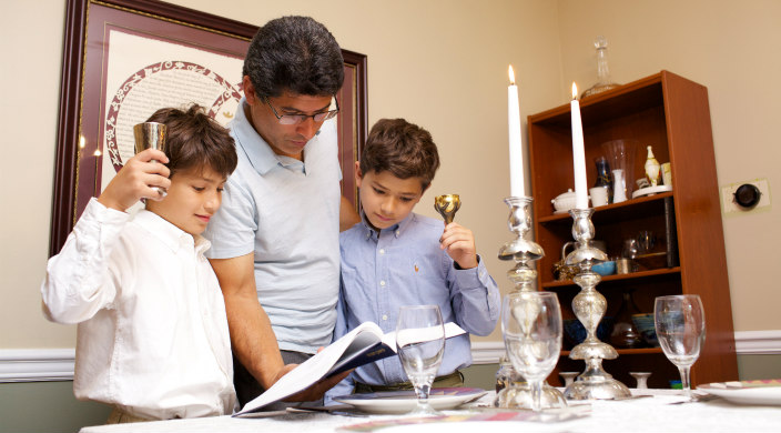 Father and young sons reading from a prayerbook as the light Shabbat candles at a kitchen table 