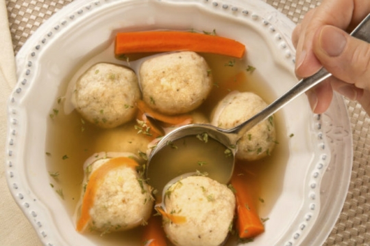Matzah ball soup in a white bowl with someones hand dipping a spoon in