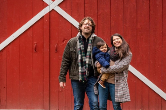 Sam and Lauren Trohman and their young son Ira pose for a family photo in front of a red and white barn