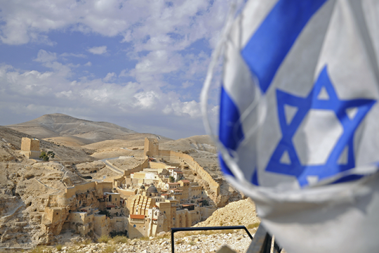 an image of  an Israeli flag with small town and mountains in the background. 