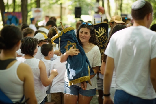 A white woman with curly brown hair and dressed in white carries a Torah with a blue cover in the midst of young campers wearing white shirts.