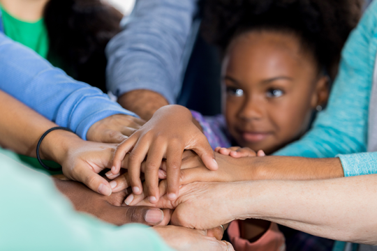 Photo of a young girl of color with her hand on top of a pile of hands in the middle of a circle
