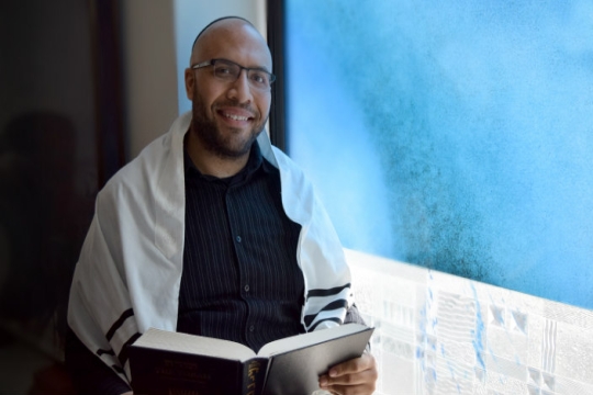 A young black man wearing a prayer shawl and a head covering while reading from a Jewish prayer book