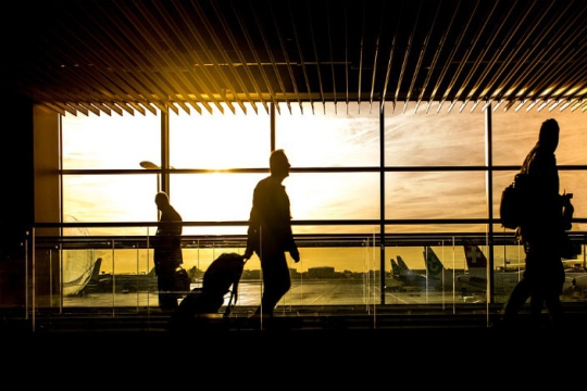 Silhouette of people carrying luggage through an airport with p;lanes out the window behind them