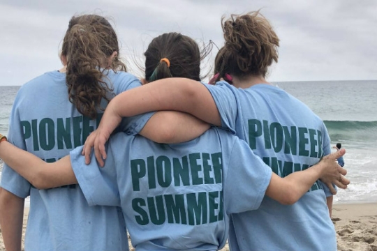 Three people facing a body of water with their arms around one another in shirts that say PIONEER SUMMER