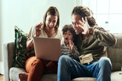 A smiling couple and their young son waving happily at a computer screen while sitting together on a couch inside a home